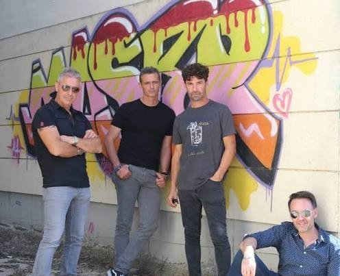 Tres miembros de Los Cucas de pie y uno sentado ante un grafiti de colores