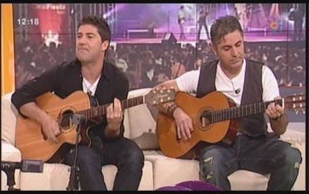 Dos miembros de Los Cucas tocando guitarras acústicas en un sofá de plató